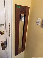 Vertical wooden framed mirror with decorative carving hung on the wall near door and light switch.
