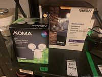 Photo showing a boxed Noma LED motion sensor security light and an Ellicott wall lantern box placed on a shelf in a basement.