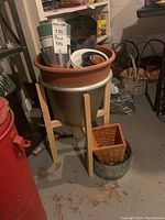 Photo capturing the large metal pot on wooden stand filled with various smaller plastic and terracotta pots, along with woven baskets in and around the main pot.