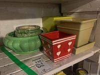 Shelf with five ceramic plant pots of various shapes and colors: large green pot with geometric design, smaller green pot with swirl detail, red square pot with white heart patterns, beige square pot with saucer, yellow rectangular pot.