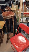 Photo showing the wooden plant stand with round top and lower shelf, and a red plastic folding chair beside it in a cluttered basement environment.