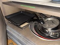 Stacked kitchen cookware including stainless steel pots with lids, black non-stick fry pans, and a metal rack.