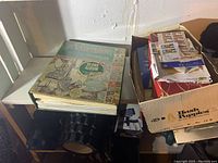 Green illustrated Le Voyageur blank album atop a stack of loose album pages in a cardboard Hush Puppies box