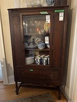 Front view of the vintage wooden display cabinet with a single glass door, inside storage shelves visible with ceramic items inside, bottom dovetailed drawer closed. Carved decorative elements on door and drawer fronts visible.