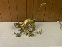 Four brass cat figurines on a white surface in front of a wood-paneled wall. Largest cat is about 14 inches long with raised tail. Two medium-sized cats and one small cat included.