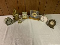 Group photo showing all seven clocks arranged in a line on a white cloth with a wood-paneled background.