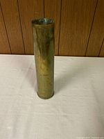 Full view of the brass cylindrical shell casing standing upright on a white surface against a wood panel background.
