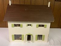Front view of the handmade wooden doll house with brown roof and green window shutters on white walls.