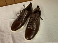 Front and top angled view of brown leather sneakers showing lace-up style, metal eyelets, and white rubber sole.