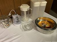 Photo showing assorted kitchen items on table including 3 springform pans, juice jug, storage containers, chopper, citrus juicer, and sea glass jar