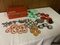 Full layout of the vintage children's play dishes including an orange carrying case, metal dishes with rose patterns, metal cupcake pans, plastic teapot and cups, metal cookie cutters, and decorative plates.