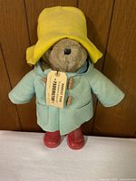 Front view of Paddington Bear standing upright showing yellow hat, blue coat with toggles, red boots and face with tag attached.