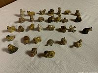 Overview of 34 miniature Wade ceramic animal figurines arranged in rows on white fabric. Animals include dogs, rabbits, birds, bears and others in brown, white, and tan glazes.