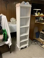 Tall white wooden shelving unit against wood paneled wall in room. Shows full front view of shelf, highlighting five shelves, decorative top with curved cutouts, and four legs with cutout detail.