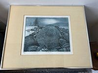 Framed etching showing a rocky winter landscape with a large boulder, signed and numbered bottom corners, displayed against a white wall.