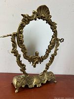 Front view of brass Rococo Revival figural cherub swing mirror on wooden surface, showing mirror frame details and cherub figures.