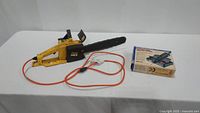 Photo showing McCulloch Electromac 14ES electric chainsaw with orange power cord and boxed Dremel tool kit on white table.