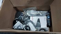 Box with assorted electronics cords, a white LG external DVD writer box, white and black cables, an RCA Universal remote on the right side, and a black flip cell phone.