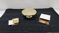 Photo of three items on black backdrop: rectangular gold wheat trinket dish with Michael Aram branding and care instructions, metallic gold-tone round cake stand, and brass 4 candle holder centerpiece