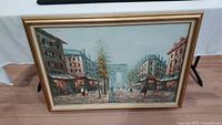 Framed oil painting depicting Paris street scene with buildings, pedestrians, trees, and Arc de Triomphe in background
