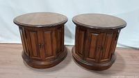 Pair of round wooden drum side tables shown from front and side angle, highlight paneled door design and base molding