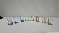 Photo showing the 8 assorted Murano glass tumblers with various colored vertical stripes lined up on a white surface.
