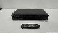 Front view of black Panasonic DVD player with disc tray and control buttons, remote control placed in front.