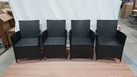 Four black rattan outdoor chairs side by side on wooden floor with visible surrounding warehouse area.