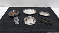 Overview photo of all six items displayed on black cloth, showing various silver plated and silver colored metal serving pieces alongside a crystal shaker.