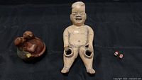 Three South American pottery figurines including a brown kissing couple, beige Olmec-style baby with a broken hand, and two tiny painted animals with gold hooks.