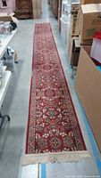 Full length view of the very long wool runner rug laid out in an aisle, showing detailed geometric floral design with red background and visible fringe at one end.