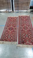 Two red wool runners laid flat on concrete floor showing geometric floral Persian style pattern with cream fringed edges.