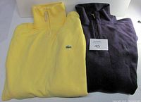 Two folded pullover sweaters stacked: purple Ralph Lauren Polo and yellow Lacoste with visible collar and embroidered logo