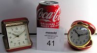 Two compact folding wind-up travel alarm clocks with red casings and gold-tone frames, displayed next to a Coca-Cola can for scale.