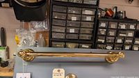 Two polished brass bath bars mounted on grey board, showing front and side views. Tags indicate brand and model. Photos show reflective golden brass finish and classic decorative mounting brackets.