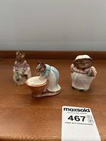 View of three Beswick Beatrix Potter ceramic figurines on wooden surface: Hunca Munca sweeping, Mr. Figgy Winkle washing, and Anna Maria.