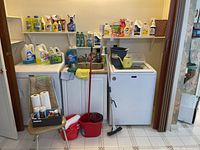 Wide view showing vintage kitchen chair, sink area, red spin bucket with mop, blue watering can, blue bucket, and cleaning products lined up on washer and dryer units.