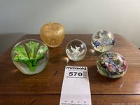 Five glass paperweights displayed on a wooden surface showing various colors and designs, including an apple shape and floral inclusions.
