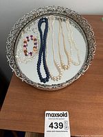 Full set of jewelry on the silver ornate mirrored dresser tray including pearl necklaces, blue bead necklace, and beaded bracelets.