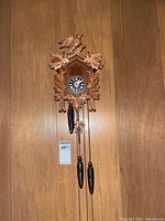 Front view of the carved wooden cuckoo clock showing bird and leaf motifs, clock face, and three pinecone-shaped weights hanging by chains.