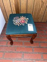 Front view of foot stool showing green needlepoint floral upholstery and curved wooden legs on brick floor.