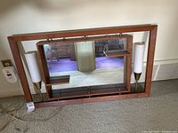 Wall mirror resting on carpet showing full wooden frame, central mirror, and two side lamps with white shades, electrical cord visible.