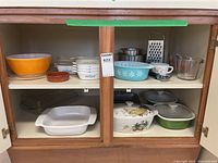 Photo showing full view of cabinet with various vintage kitchen dishes and bakeware including covered casserole dishes and white bakeware.