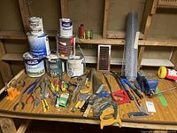 Overview of various cans of paint, hand tools including pliers, screwdrivers, saws, and a clutch of paint brushes on a wooden table.
