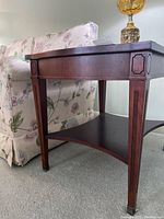 Side view of one Knechtel end table showing veneered wood surface, carved vertical leg accents, brass feet, and lower shelf
