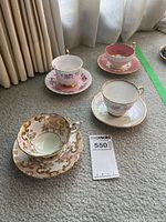 Four vintage teacups and saucers displayed on a carpeted floor with a MaxSold tag visible. Various floral and gilded gold decorative patterns visible on all pieces.