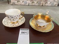 Two teacup and saucer sets on a dark wood table. One white with delicate gold floral patterns, one fully gold with turquoise circular accents.