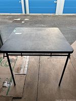 Square folding card table standing opened, showing black tabletop and metal legs.