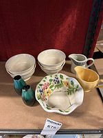 Overview of six white ceramic bowls, one yellow ceramic bowl, a small black and white striped creamer, two small blue gradient salt and pepper shakers, and one hand-painted floral ceramic bowl with scalloped edges.