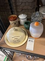 Overview of six cheese pots on shelf including large ceramic cheese dome, brown ceramic pot, white enamel pot, small green pot, blue and white speckled pot, and metal pot with wear.
